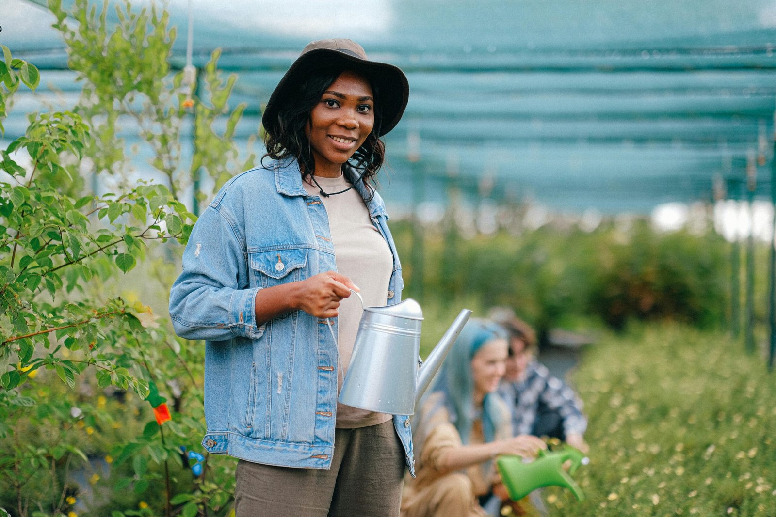 A cheerful woman holding a watering can in a greenhouse, tending to plants.