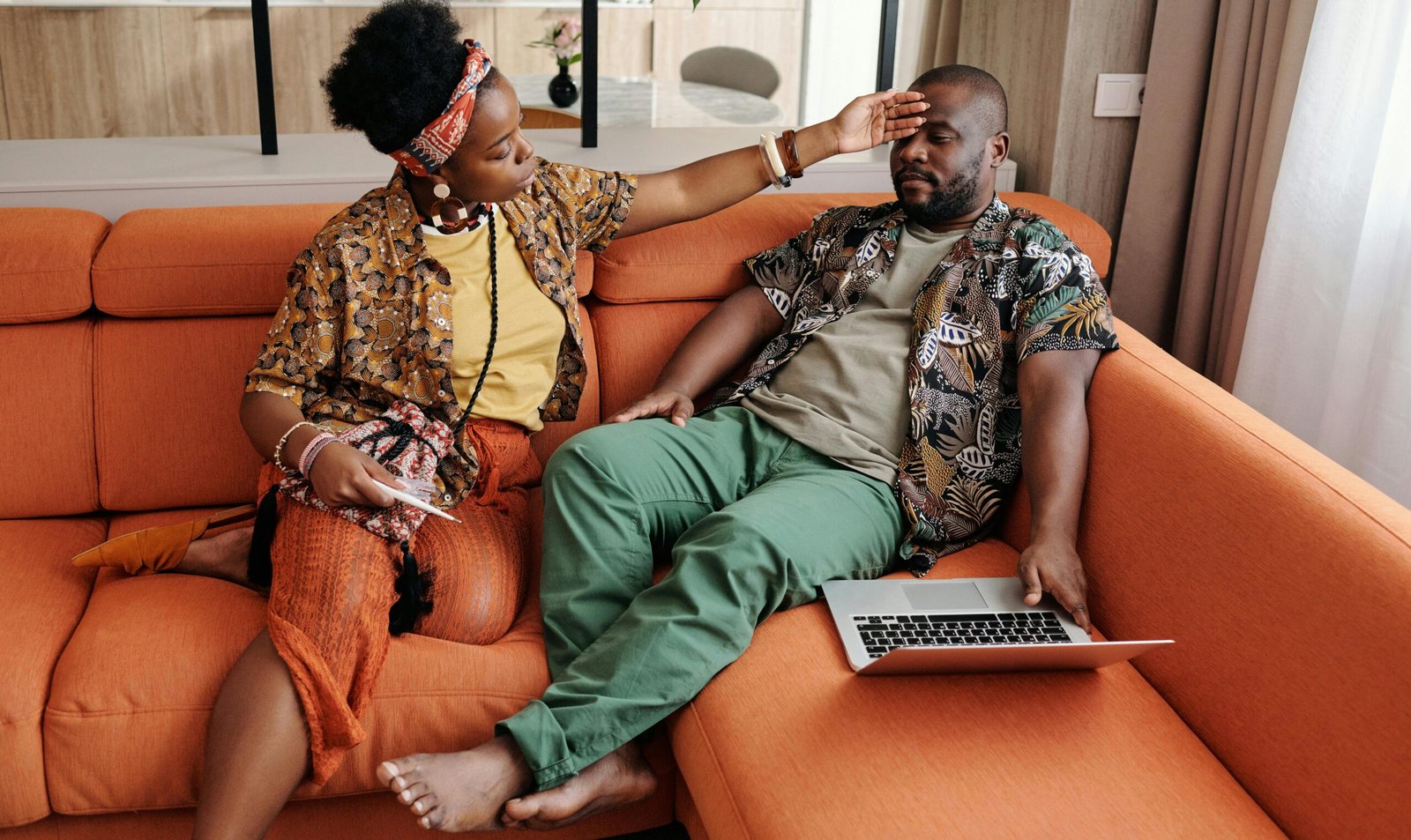 A concerned woman checks her partner's temperature as he rests on the sofa, caring at home.